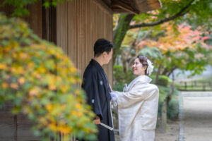 後楽園で和装姿の新郎新婦が前撮りを行う様子。紅葉と日本庭園に囲まれた穏やかな風景。