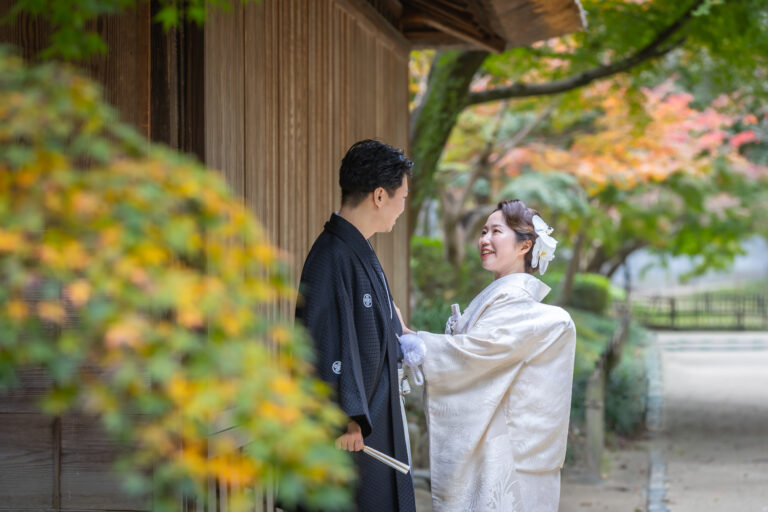 後楽園で和装姿の新郎新婦が前撮りを行う様子。紅葉と日本庭園に囲まれた穏やかな風景。
