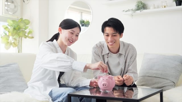 笑顔で貯金箱にお金を入れる夫婦の姿。未来への希望と家族の絆を象徴。