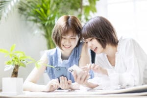 スマホを見ながら笑顔で話す20代女性二人｜岡山の婚活で情報収集するイメージ